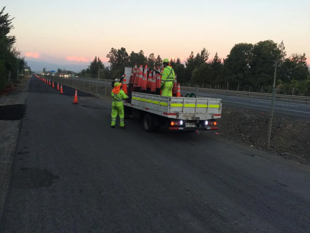 Camión de la cuadrilla junto a un operario con ropa reflectante instalando conos en la carretera al atardecer.