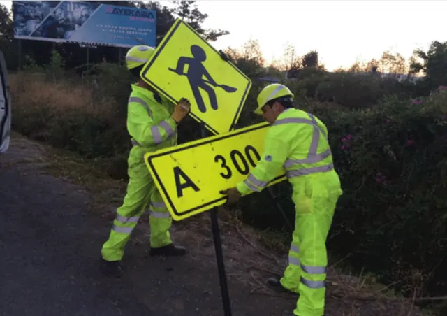 Dos operarios con ropa reflectante instalando una señal vial de cruce peatonal a la orilla del camino.